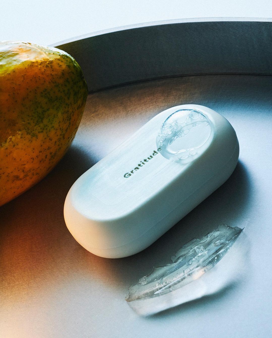 White soap bar with 'Gratitude' text sits on a silver tray next to an orange and translucent soap piece in a studio shot.