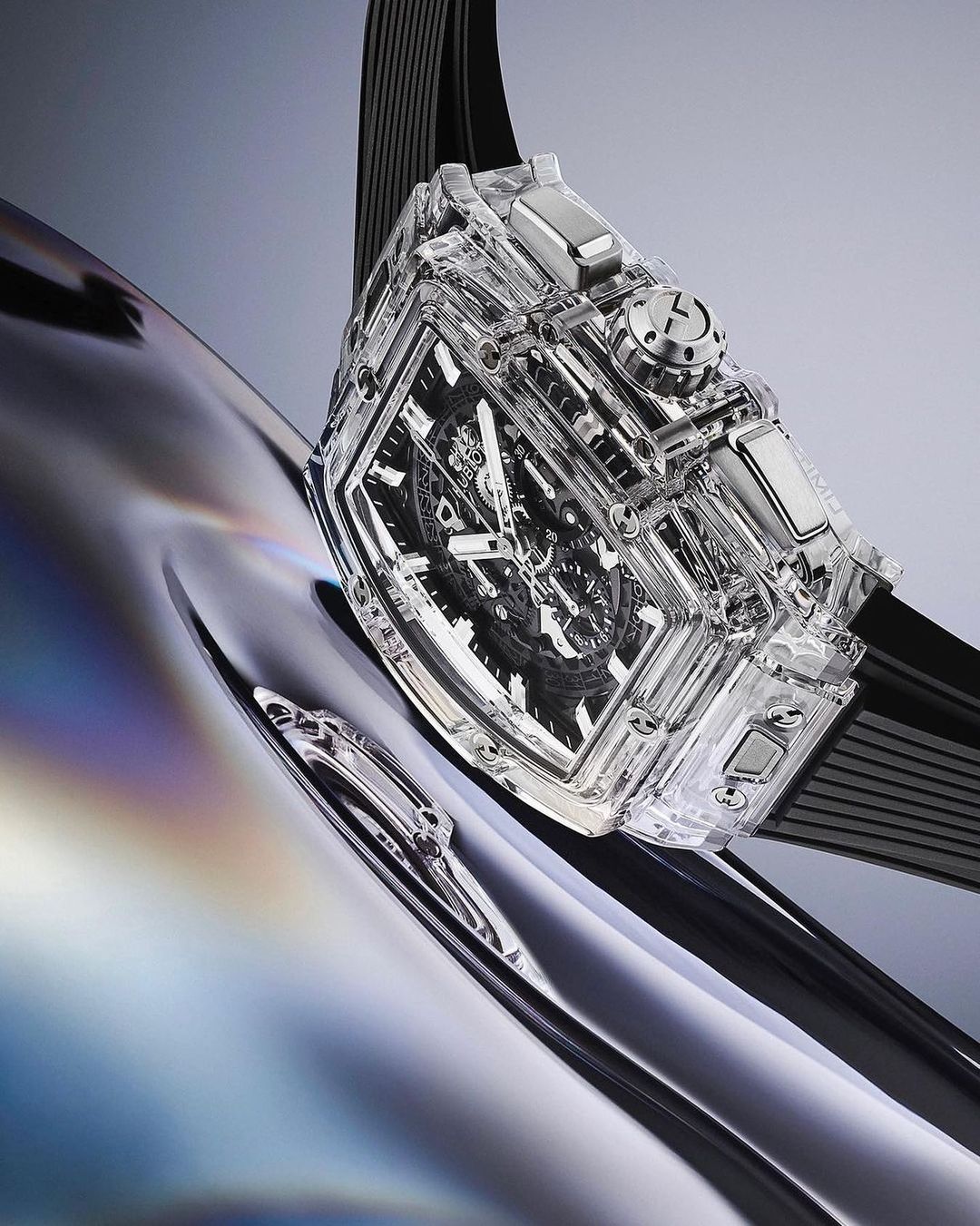 Close-up of a clear-encased mechanical watch with black strap on a reflective, metallic silver surface, emphasizing luxury and craftsmanship.