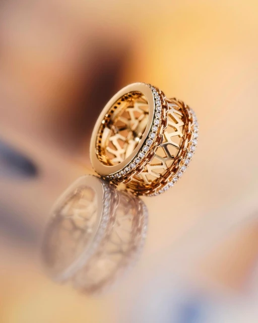 Close-up of a gold statement ring with intricate geometric patterns and diamond accents, reflecting on a soft, gradient background.