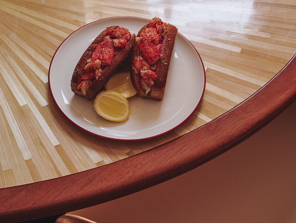 Two lobster rolls with lemon wedges served on a white plate with red trim, set on a wooden table.