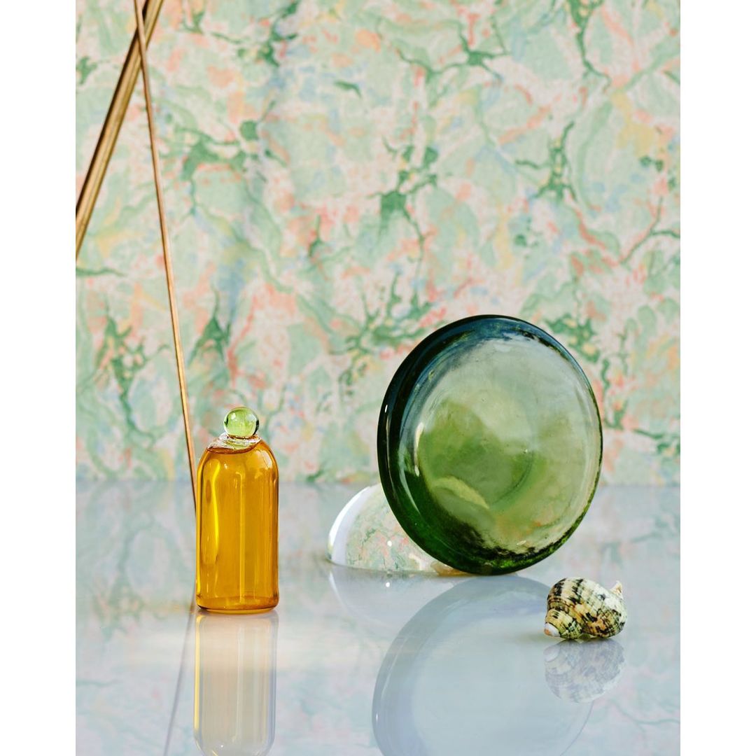 A still life featuring a golden oil bottle, a transparent orb, and a shell on a reflective surface, set against a patterned backdrop.