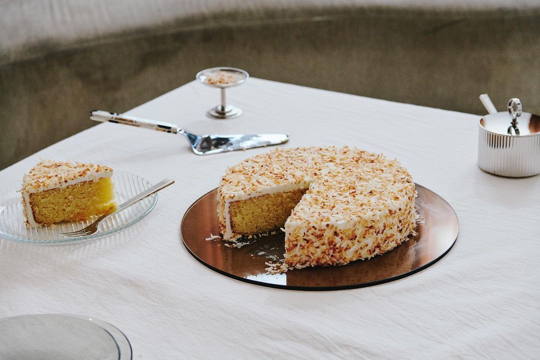 Coconut cake with a slice cut out sits on a copper plate, presented with a cake server and a slice on a clear glass plate.