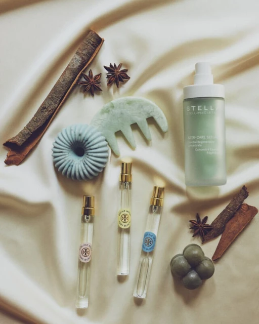 Still life composition on a silk cloth featuring perfume bottles, a hair serum, soap, gua sha comb, cinnamon sticks, and star anise.