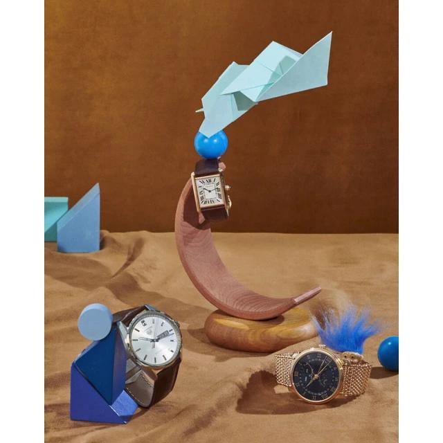 Three wristwatches displayed on brown and tan surface with blue, geometric objects and a light blue paper airplane on a brown background.