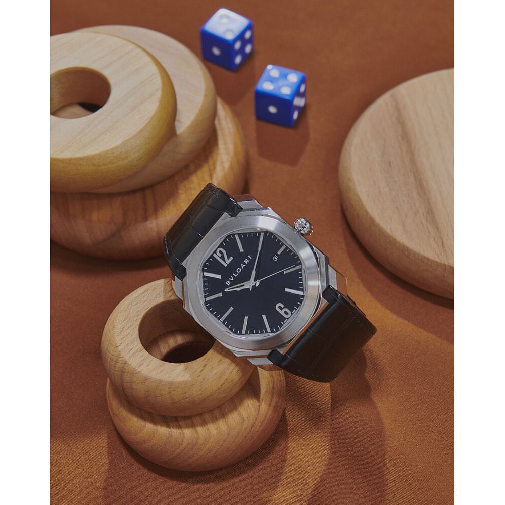 Luxury wristwatch with a silver case and black leather strap displayed on a brown surface with wooden toys and blue dice.