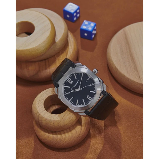 Luxury wristwatch with a silver case and black leather strap displayed on a brown surface with wooden toys and blue dice.