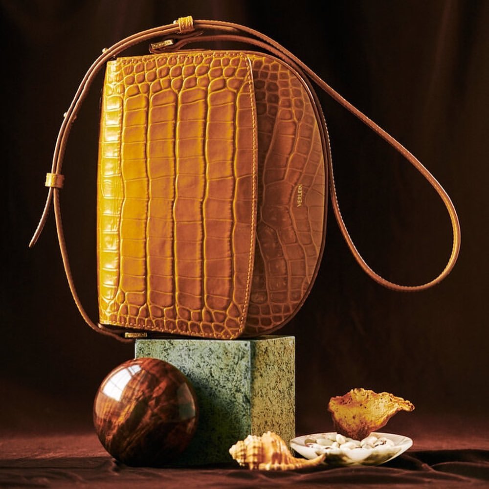 Designer crocodile-texture handbag in shades of brown and gold is displayed with decorative objects against a dark brown background.