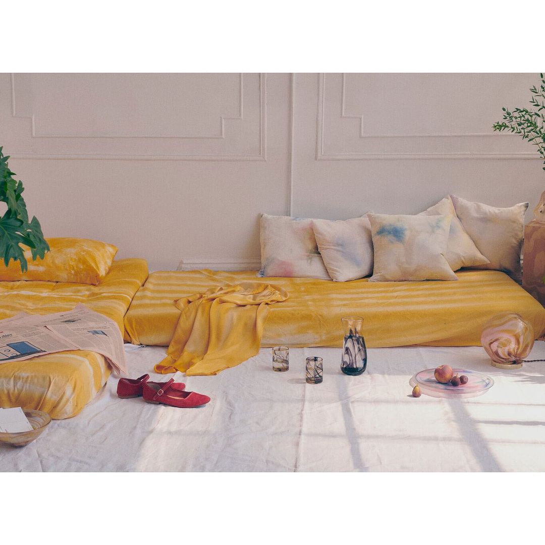 Still life composition features a yellow sofa, decorative pillows, glassware, a newspaper, and red Mary Jane shoes in soft natural light.
