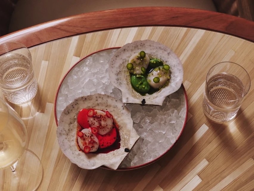 Overhead shot of two scallop shells filled with seafood, served on ice on a wooden table, accompanied by a glass of white wine and water.