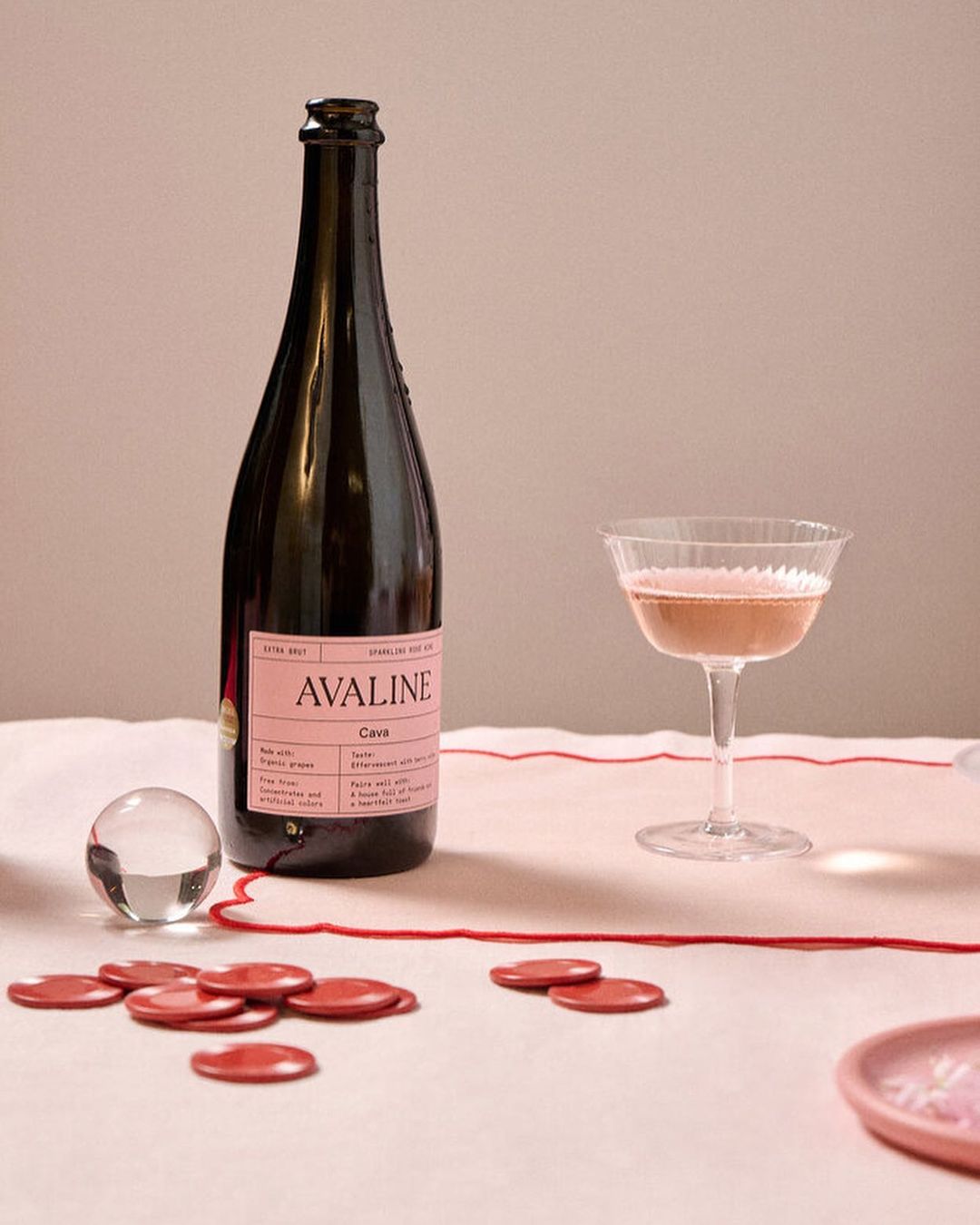 Elegant still life featuring a bottle of Avaline cava alongside a stemmed glass with bubbly rosé wine, red wafer seals, and a glass orb.