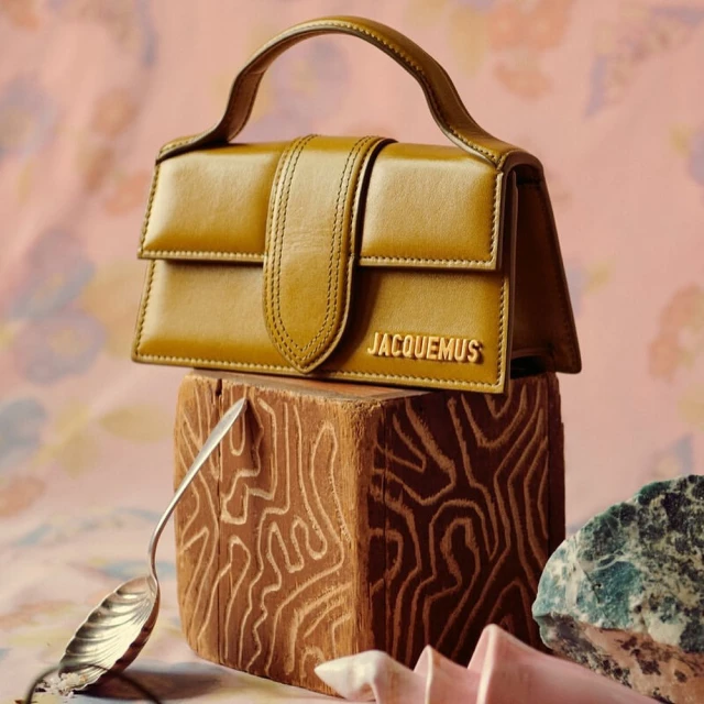 Jacquemus Le Chiquito handbag in mustard leather displayed on a patterned backdrop with a textured wooden block and raw stone.