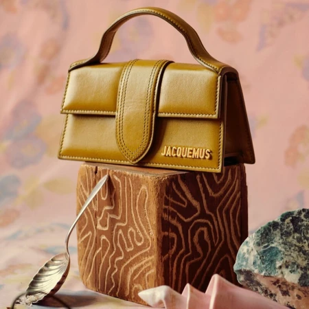 Jacquemus Le Chiquito handbag in mustard leather displayed on a patterned backdrop with a textured wooden block and raw stone.