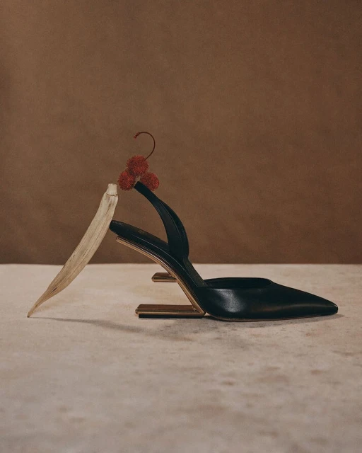 Studio shot of a black slingback heel with unique architectural gold heel, a neutral leaf and red berries, on a textured surface.