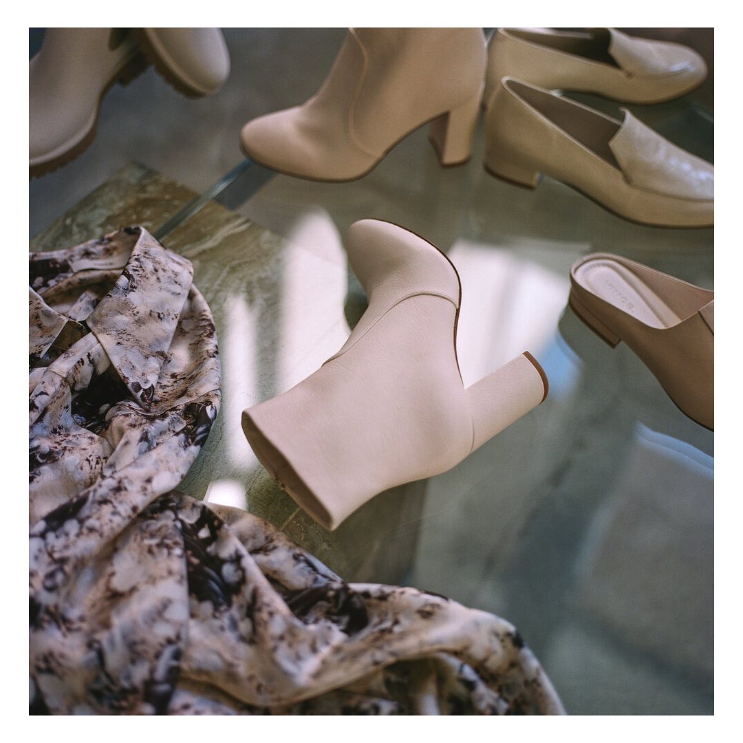 Beige ankle boots and pumps are showcased on a marble surface, accompanied by a patterned blouse, creating a fashion-forward aesthetic.