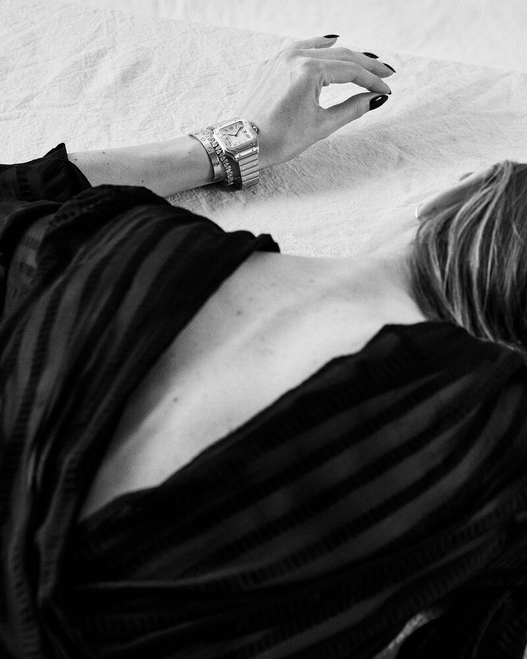 Woman with black nail polish lies on a white surface wearing an ornate metal watch and bracelets; black and white medium shot.