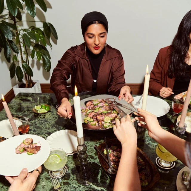A woman in a headscarf dines with a group at a table laden with plates, cocktails, and candle light, fostering a warm, inviting atmosphere.