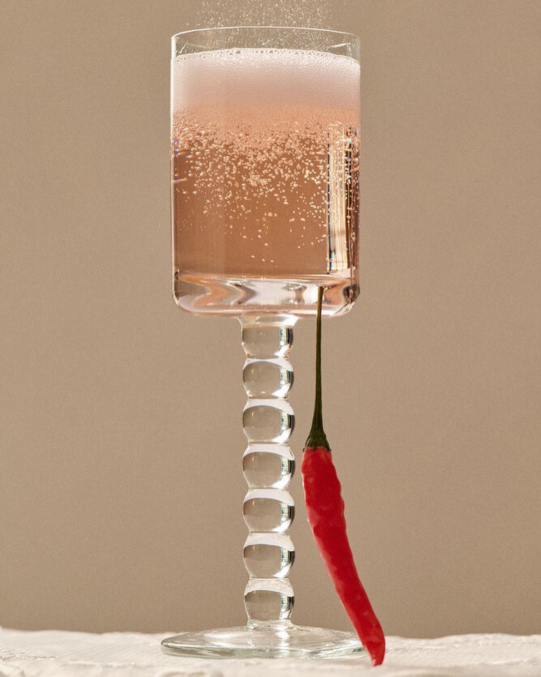 Tall glass of sparkling pink cocktail with bright red chili pepper garnish against a neutral background on a white tablecloth.