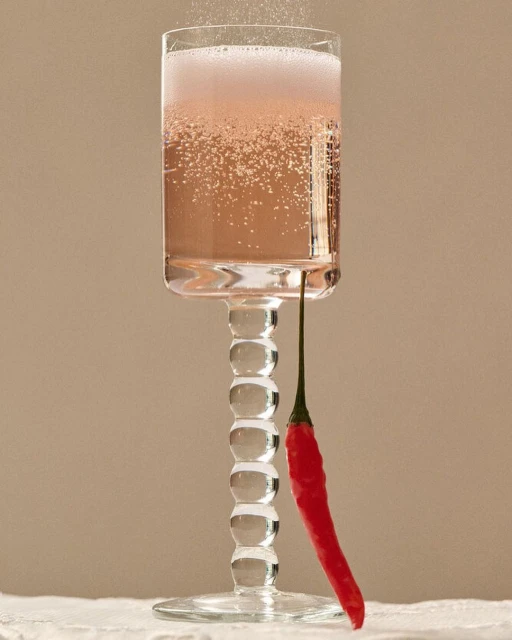 Tall glass of sparkling pink cocktail with bright red chili pepper garnish against a neutral background on a white tablecloth.