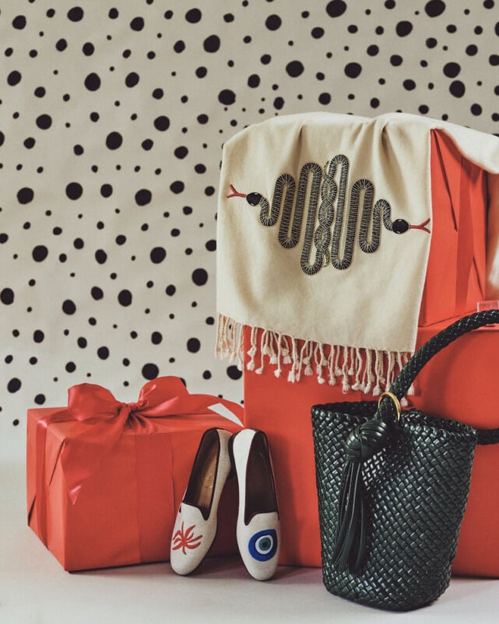 Still life with wrapped red gifts, patterned scarf with snake embroidery, black tote bag, and decorative slip-on shoes on a white surface.