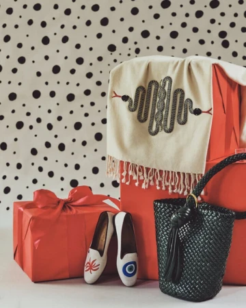 Still life with wrapped red gifts, patterned scarf with snake embroidery, black tote bag, and decorative slip-on shoes on a white surface.