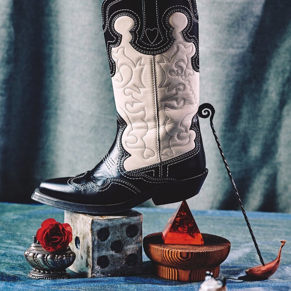 Fashion still life features a black and white cowboy boot atop a dice cube, a wood stand with a red resin pyramid, and metal ornaments.