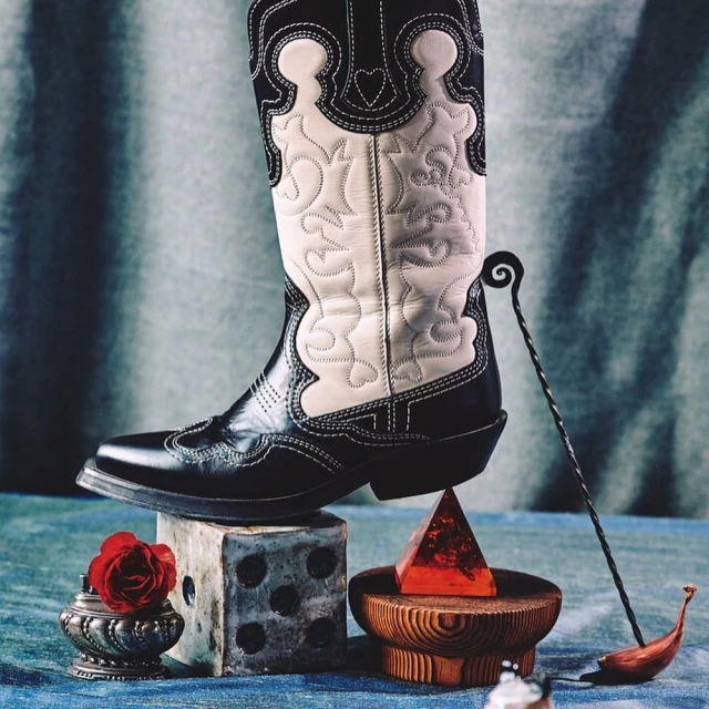 Fashion still life features a black and white cowboy boot atop a dice cube, a wood stand with a red resin pyramid, and metal ornaments.