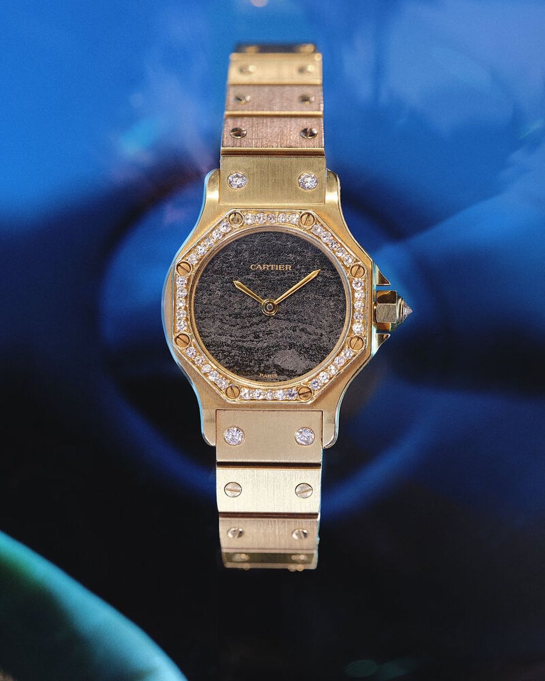 Elegant gold Cartier watch with diamonds around the face on a blue background, showcasing luxury timepiece design and craftsmanship.