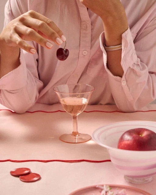 Person holding a cherry over a wine glass with pink drink, red apple, and pink tableware on a light pink tablecloth indoors.