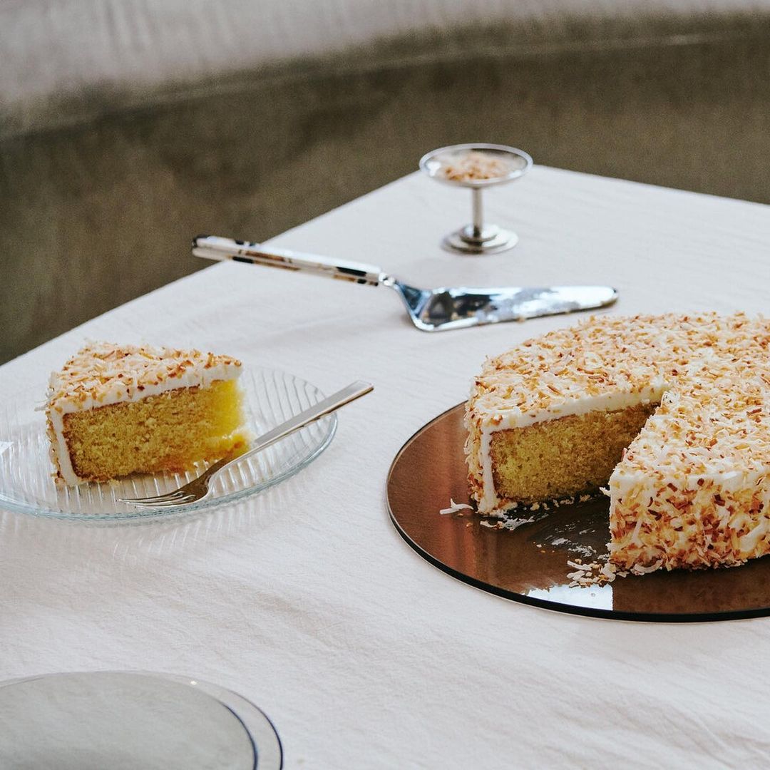 Overhead shot of coconut cake on a white tablecloth, with a slice on a glass plate, server, and dessert dish in a brightly lit indoor setting.