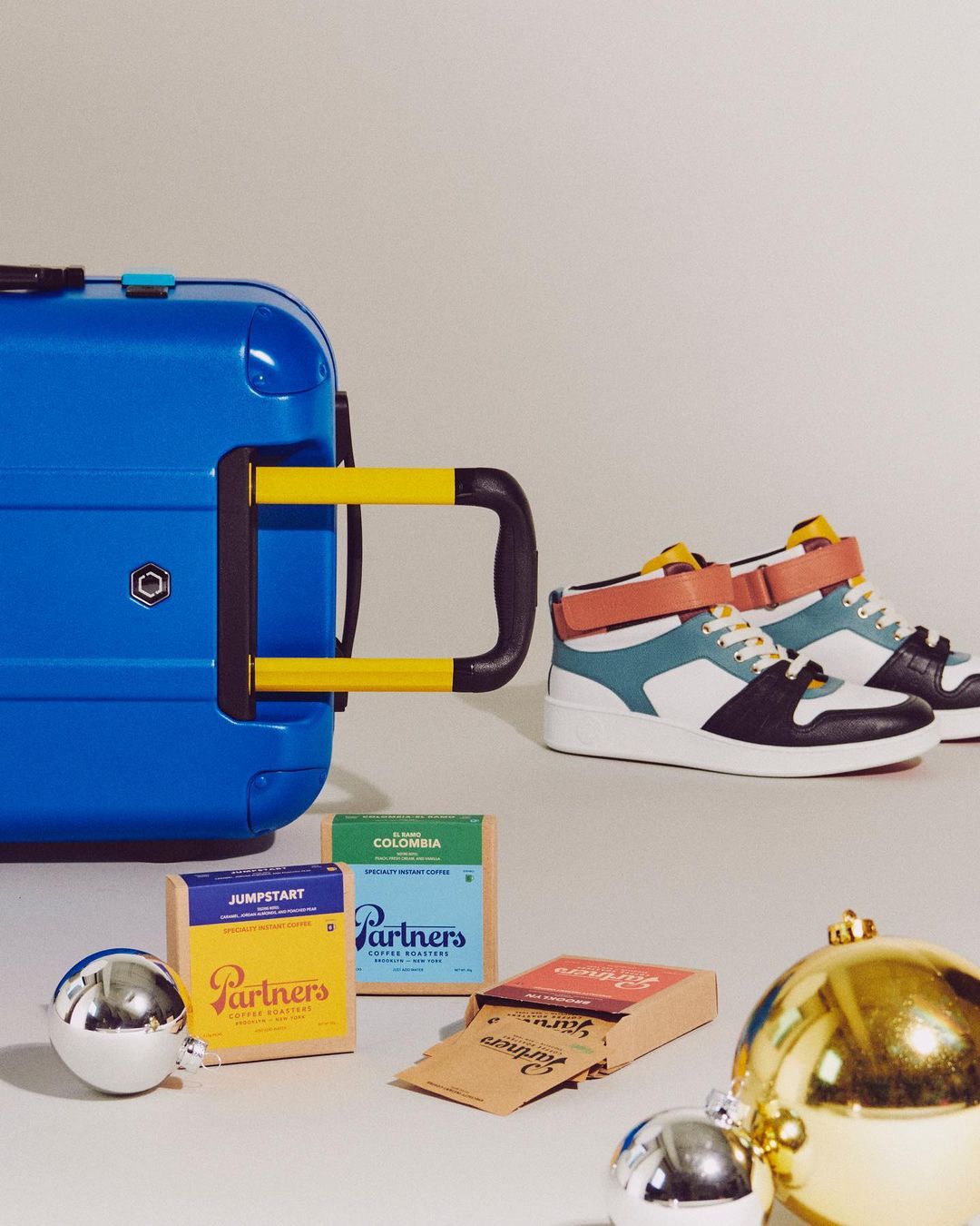 Still life shot of a blue suitcase next to colorful sneakers, boxes of Partner's coffee, and shiny silver and gold Christmas ornaments.