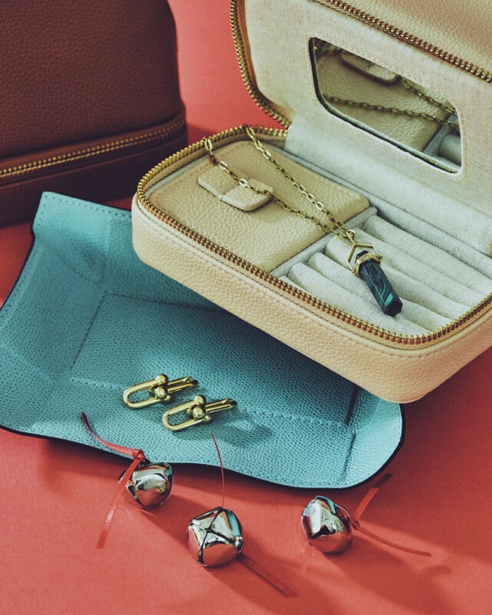 Beige jewelry case with gold chain necklace, matching earrings, silver bells, and light blue jewelry mat on a coral surface.
