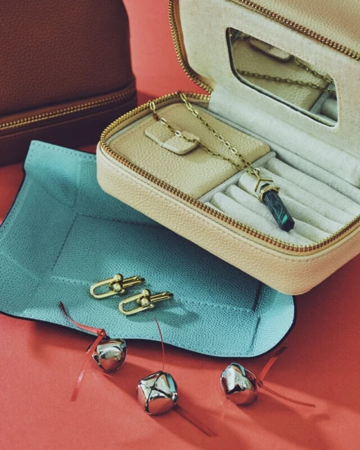 Beige jewelry case with gold chain necklace, matching earrings, silver bells, and light blue jewelry mat on a coral surface.