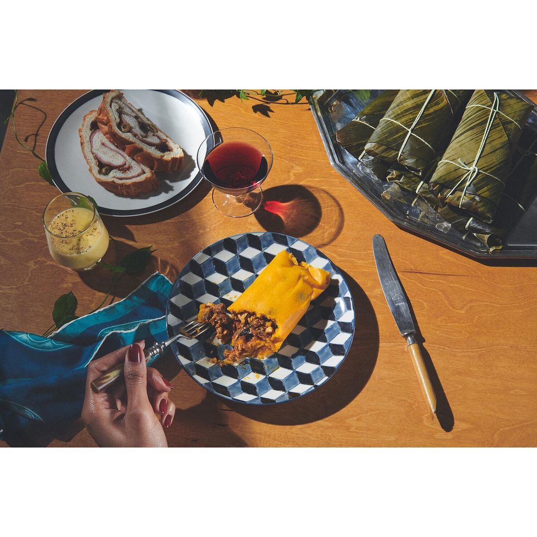 Hallacas on a patterned plate with a hand holding a fork, paired with wine and relleno, on a wooden table in warm light.