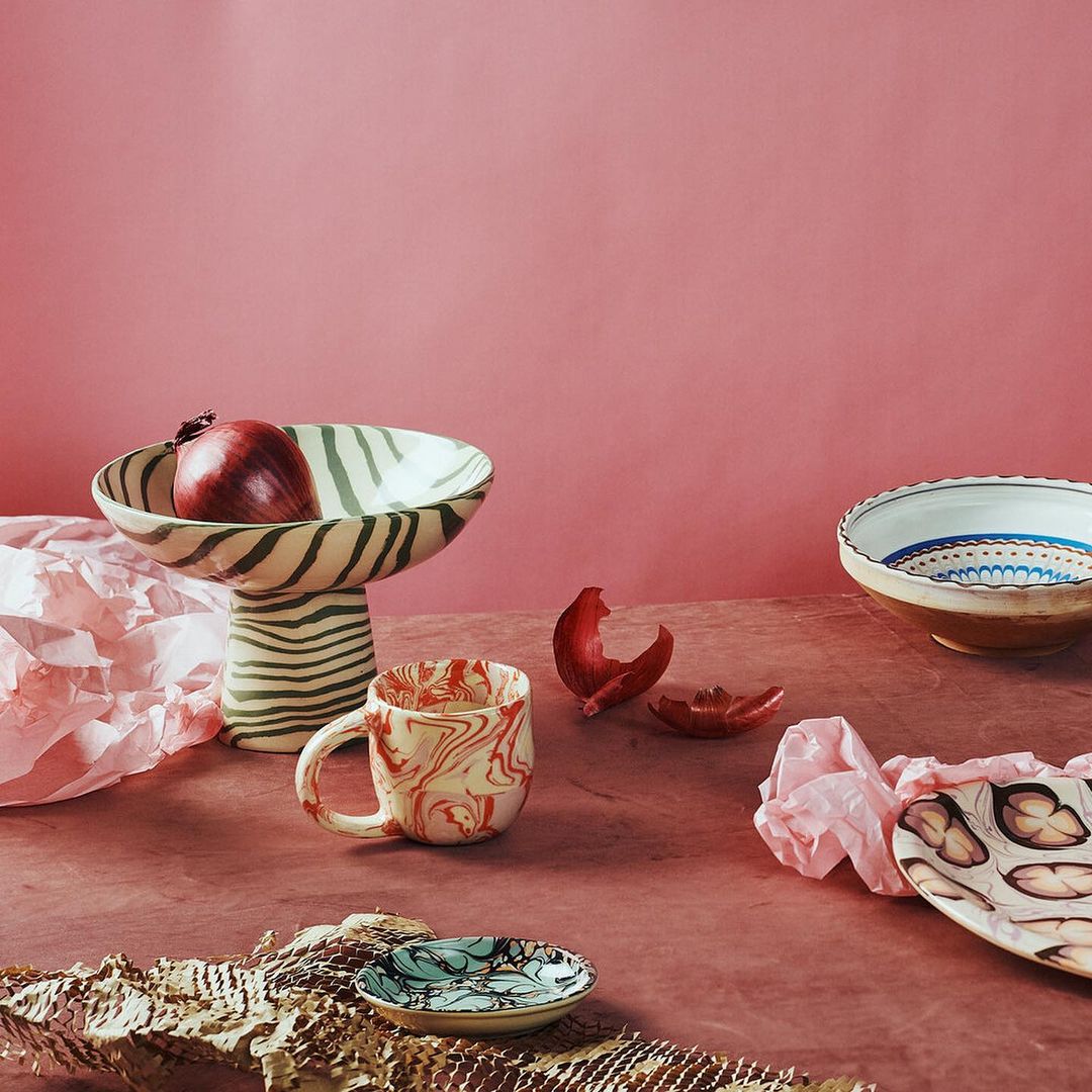 Still life studio shot with ceramic dishware in muted colors. A bowl with an onion, a mug, and small plates sit on a pink background.