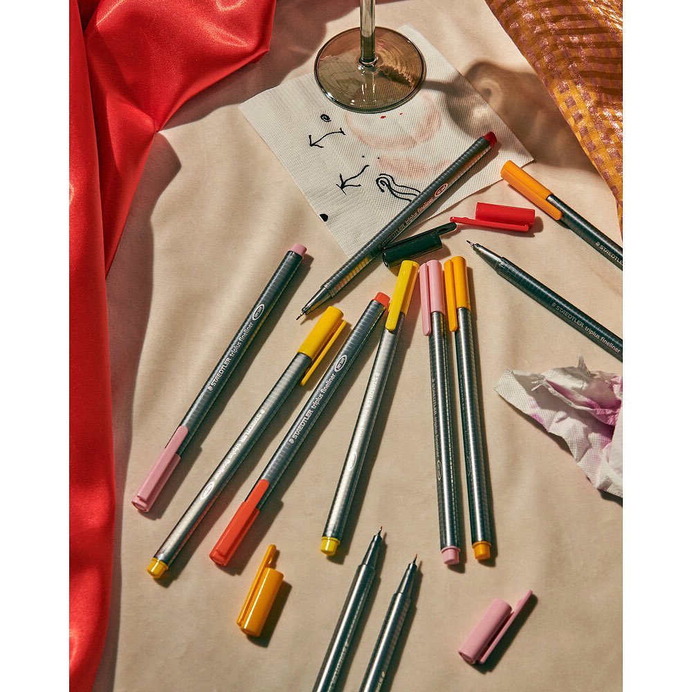 Overhead shot of colorful fineliner pens scattered on a surface with a wine glass, a napkin, and red and gold fabrics.