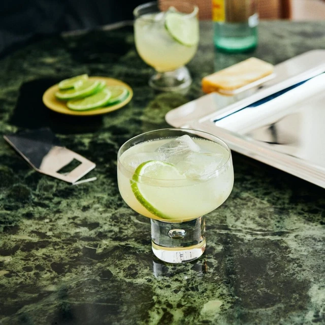Close-up of two cocktail glasses with lime slices and ice, accompanied by a serving tray and a yellow plate with lime wedges.
