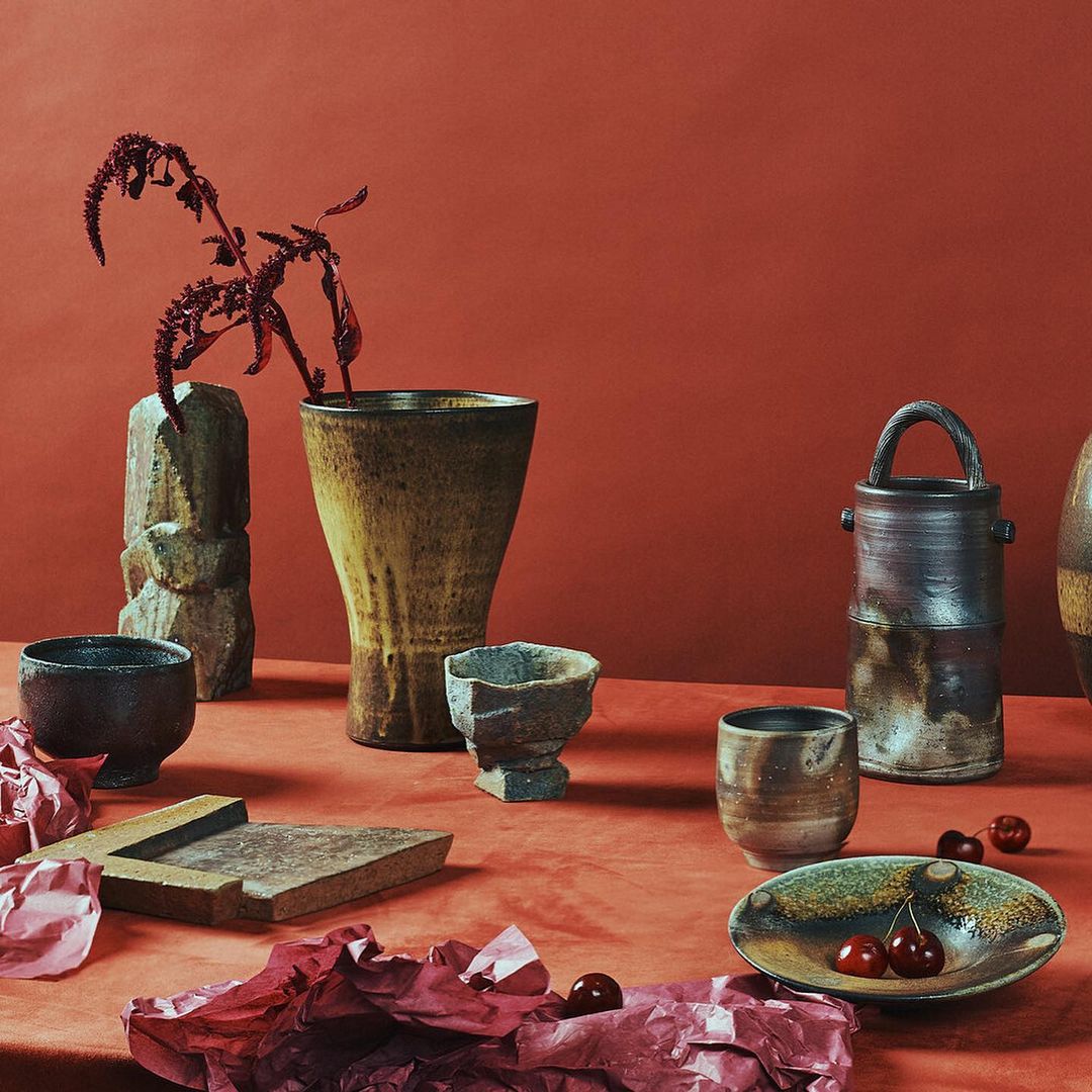 Still life studio photograph of a collection of artisan ceramic vases, bowls, and plates arranged on a rust-colored surface with dried florals and cherries.