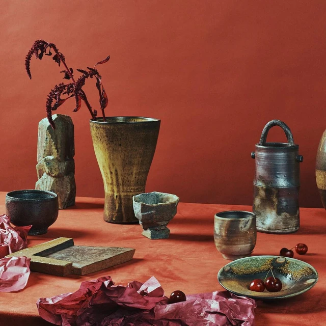 Still life studio photograph of a collection of artisan ceramic vases, bowls, and plates arranged on a rust-colored surface with dried florals and cherries.