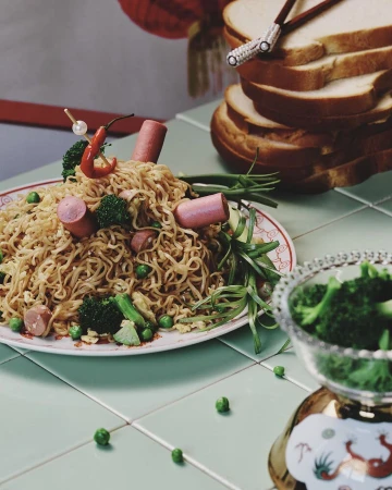 Overhead shot of noodle dish with sausage, broccoli, peas and red pepper, resting on tiled surface, with stack of bread.