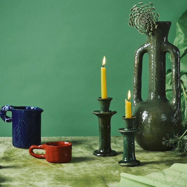 Still life with ceramic vases, candle holders with lit candles, against a green backdrop. The arrangement includes a blue pitcher and red cup.