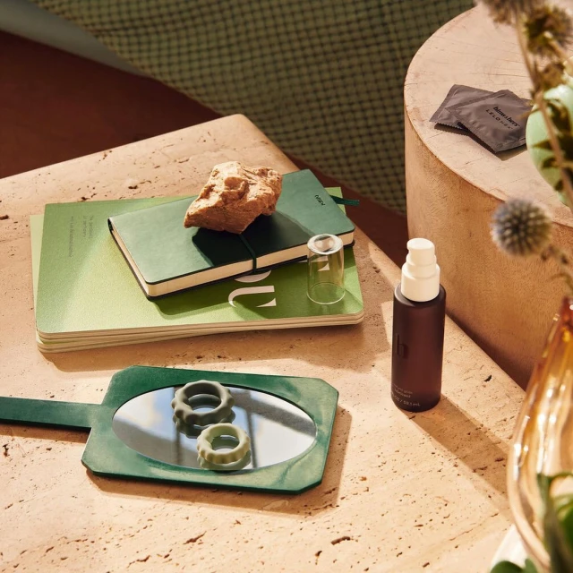 Still life on a table with green notebooks, hand mirror with rings, brown skincare bottle, rock, condom packets, and amber glass vase.