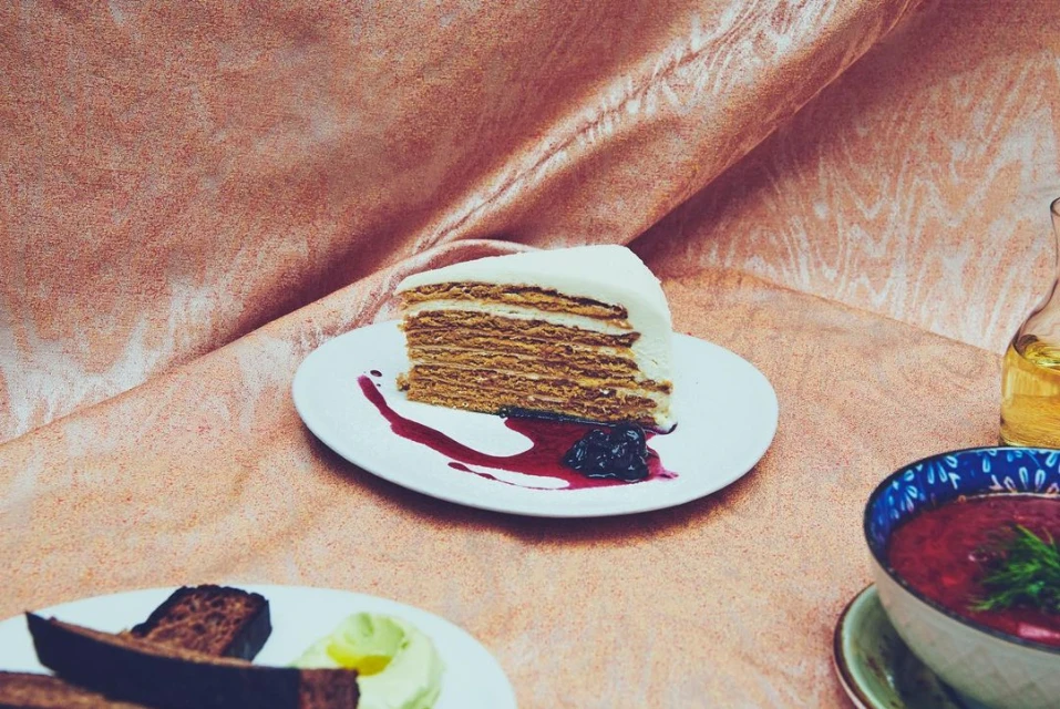 Slice of honey cake with white frosting on a white plate with berry sauce, accompanied by a bowl of beet soup and bread.