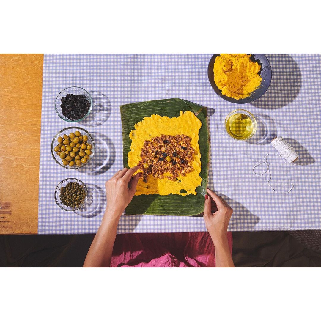 Overhead shot of a person preparing hallacas on a banana leaf with ingredients like meat, dough, olives, and spices on a checkered tablecloth.