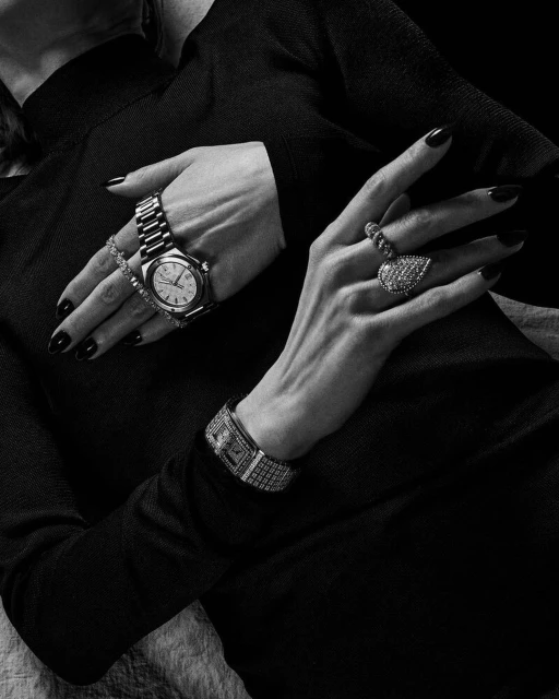 Close-up of a woman's arms adorned with a wristwatch, jeweled bracelet, and large ring, all on a textured background, in black and white.