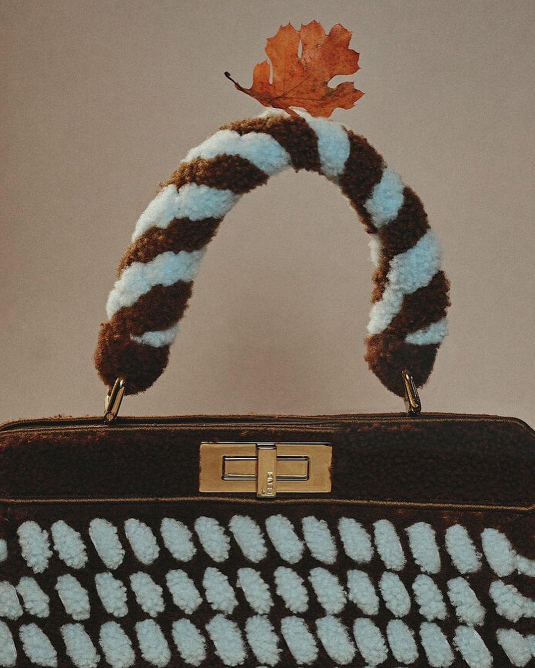 Brown handbag with a fuzzy, striped handle and a single leaf on top, set against a neutral backdrop in a studio shot.