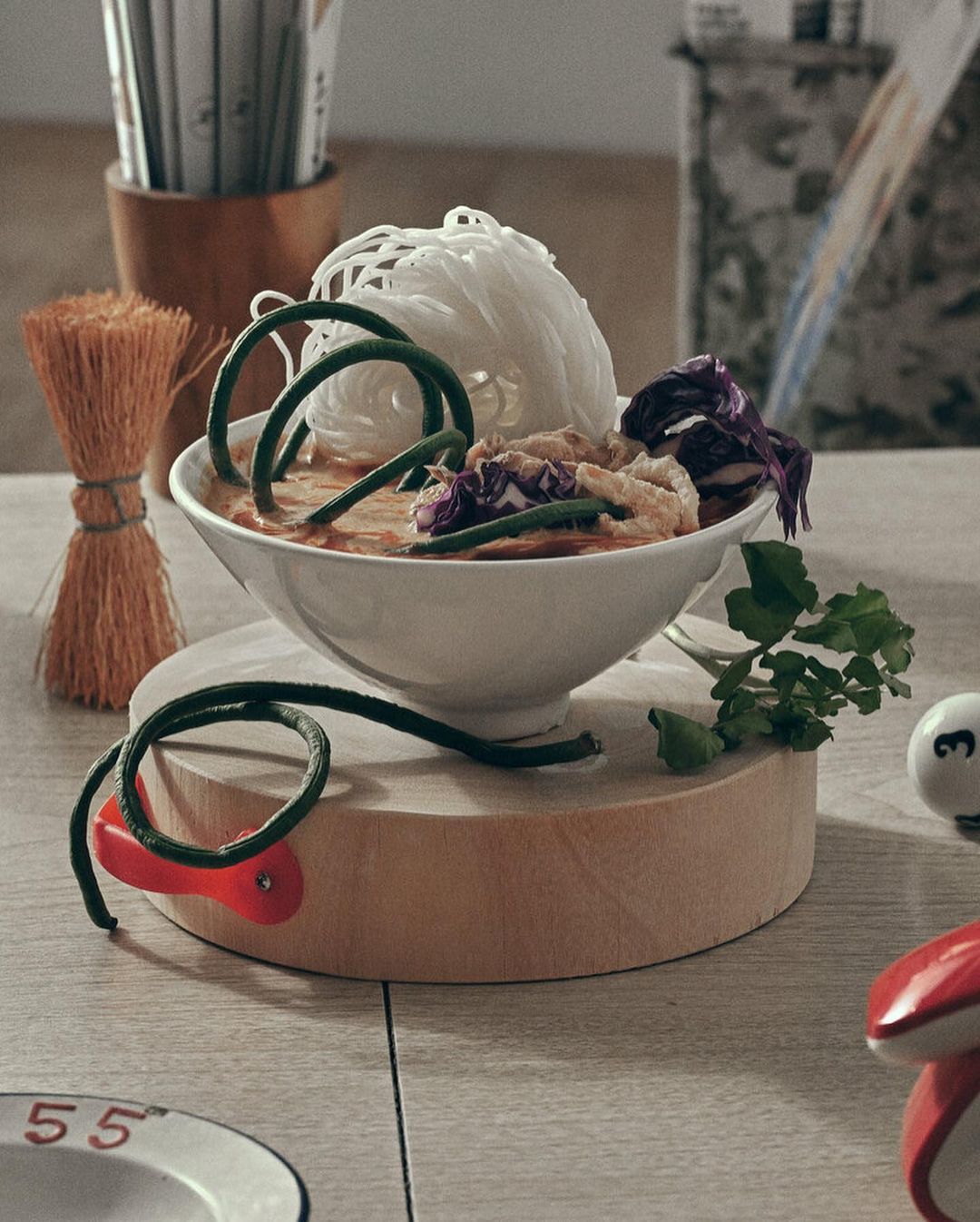 A bowl of Asian noodles with vegetables is served on a wooden stand, accompanied by chopsticks and other dining accessories on a table.
