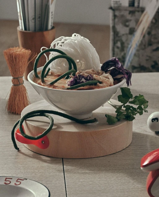 A bowl of Asian noodles with vegetables is served on a wooden stand, accompanied by chopsticks and other dining accessories on a table.
