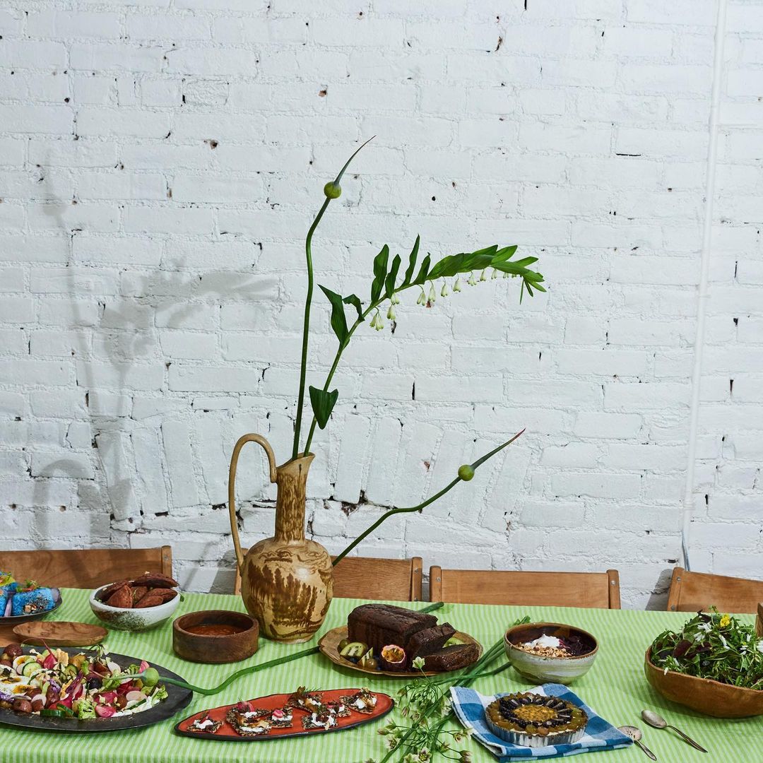 Still life of salads, bread, sauces, and dessert arranged on a green striped tablecloth with a flower arrangement and white brick background.
