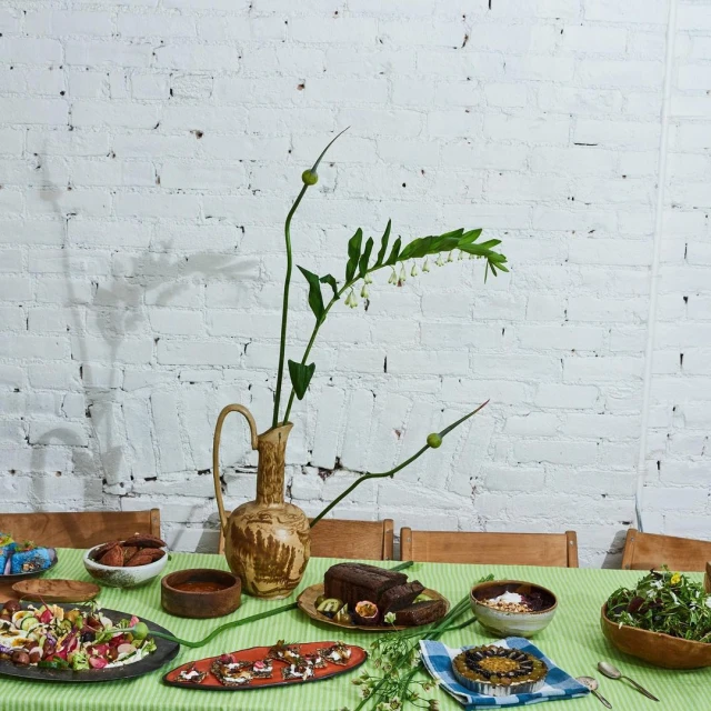 Still life of salads, bread, sauces, and dessert arranged on a green striped tablecloth with a flower arrangement and white brick background.