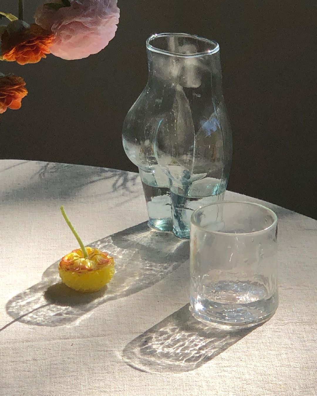 Still life composition with clear vases filled with water, a small yellow fruit, and flowers, all sitting on a textured white surface.